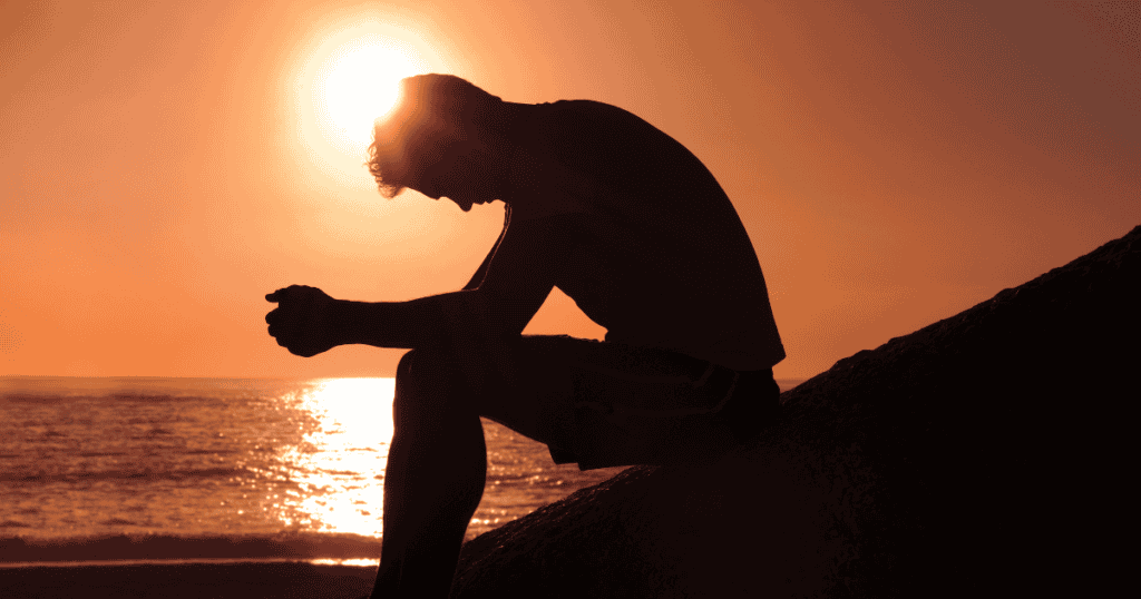 Silhouette of a person crouching and reading or adjusting something near the edge of a beach at sunset, with the sun low over the ocean.