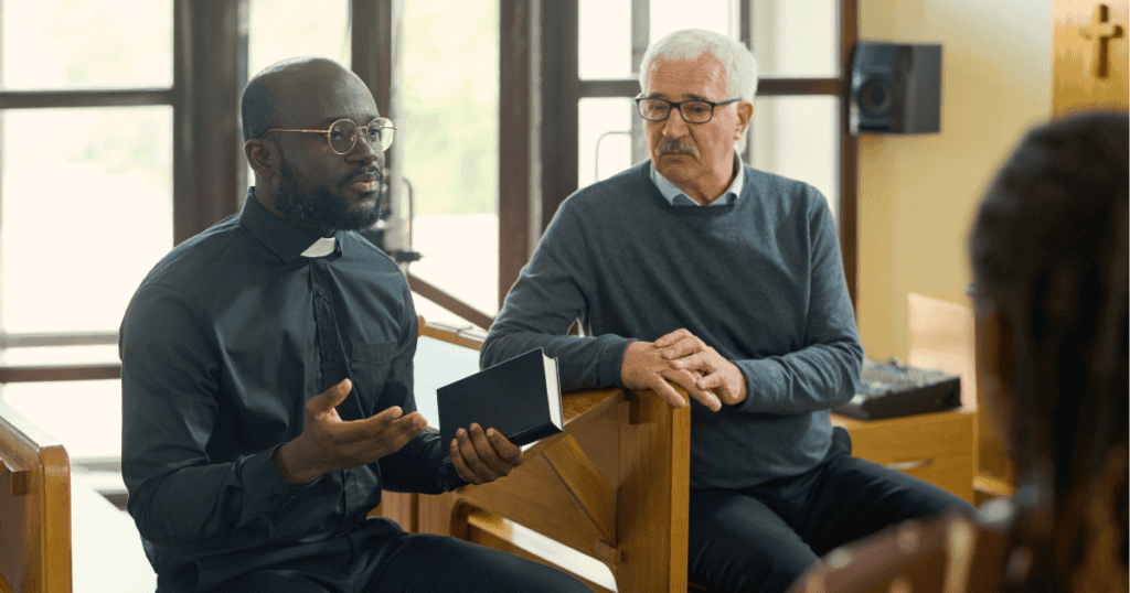 Two men in church pews talk during a service, one holding a book and the other gesturing while seated in a wooden sanctuary with large windows.